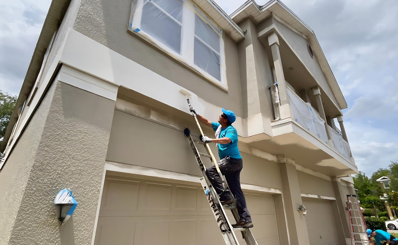 A crew member in a teal shirt stands on a ladder painting the exterior trim and stucco of a two-story house with windows taped off in plastic, with another crew member working below.